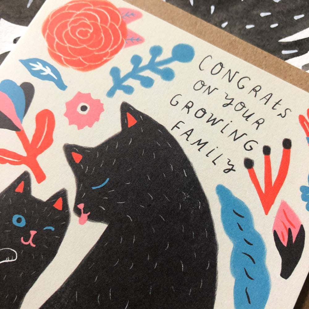 Cute greeting card featuring stylized black cats surrounded by colorful floral designs and text reading "Congrats on your growing family" for new parents or pet owners