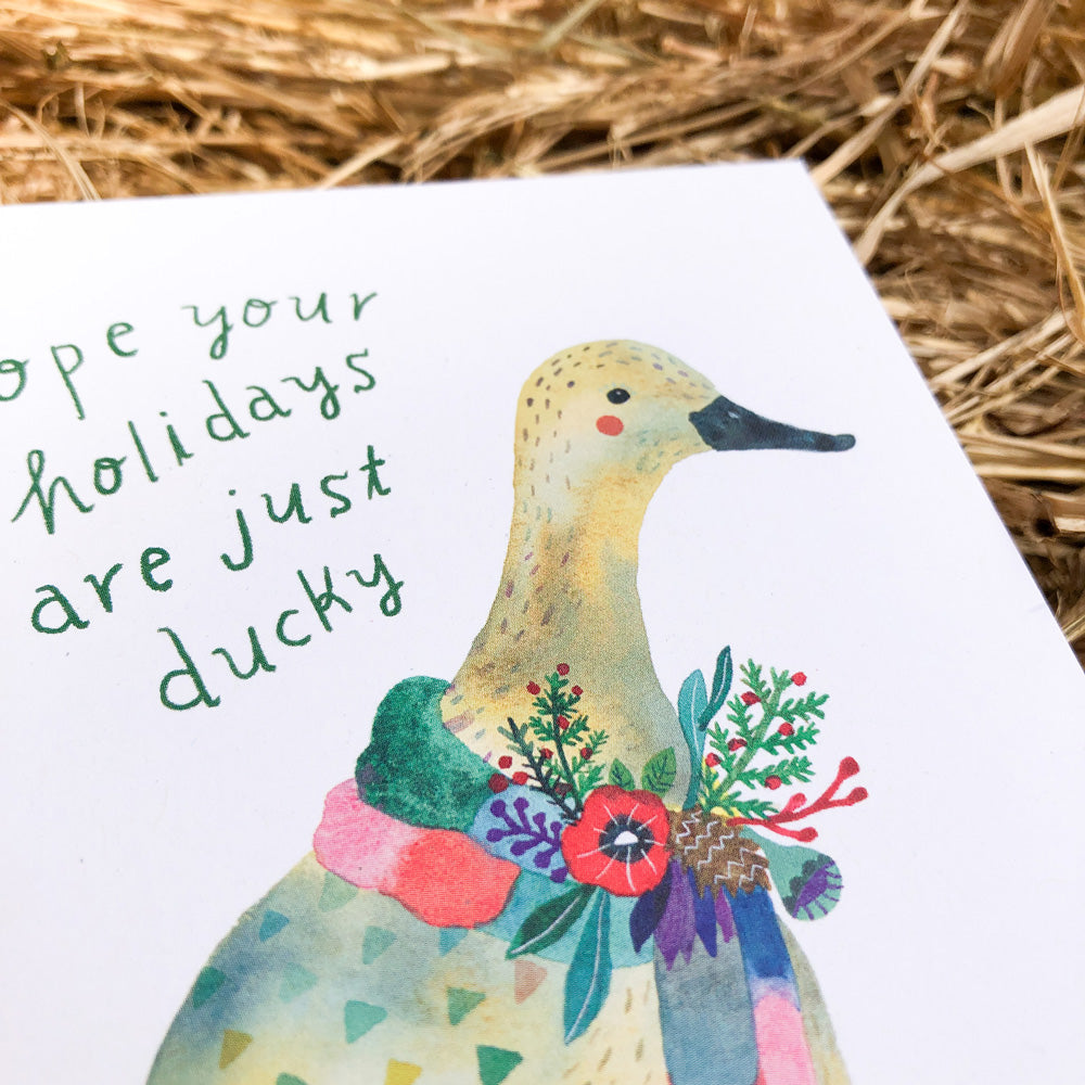 Watercolor greeting card featuring a cheerful duck adorned with colorful flowers and the playful text "Hope your holidays are just ducky" against a rustic hay background