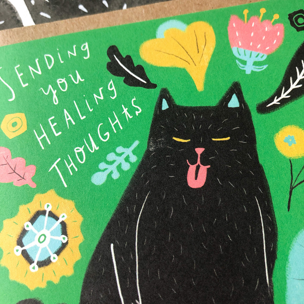 Colorful sympathy card featuring a black cat surrounded by whimsical flowers and leaves on a green background, with handwritten text "Sending you healing thoughts" in white letters