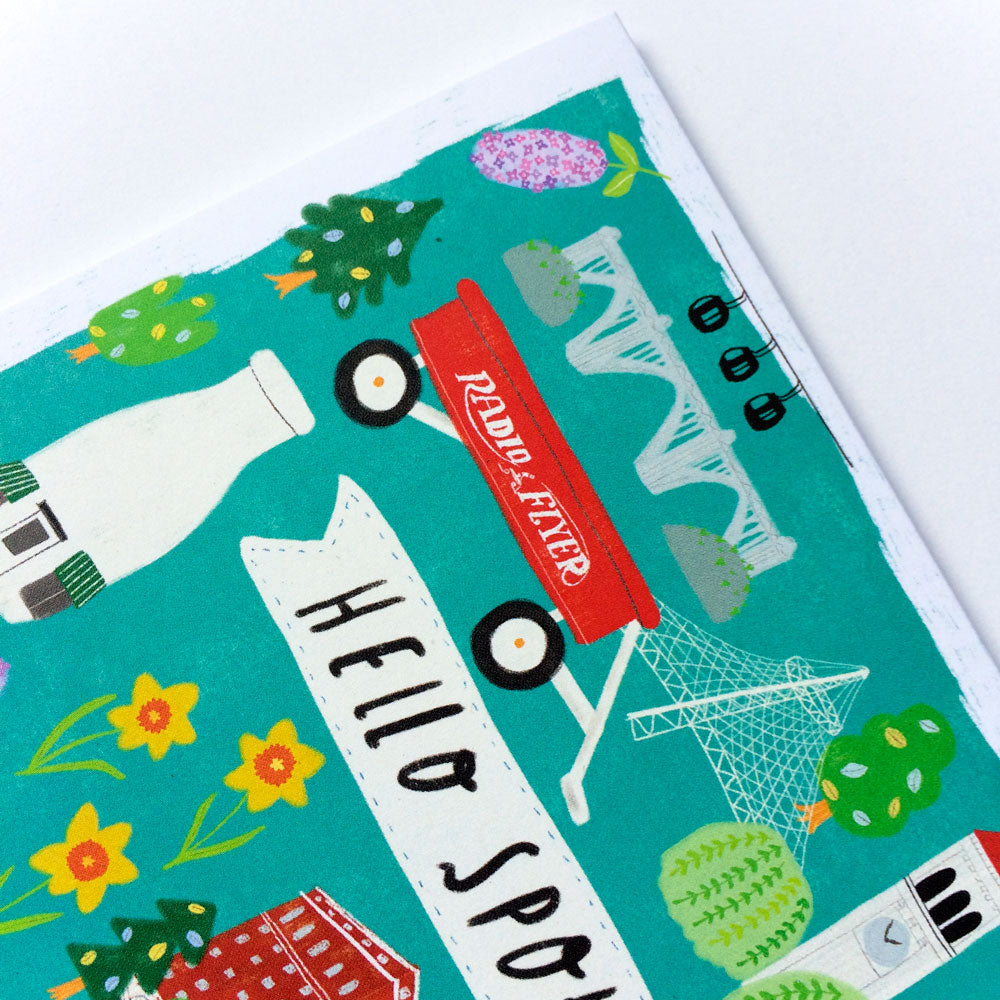 Colorful greeting card featuring iconic Spokane landmarks and symbols including a red wagon, milk bottle, bridge, and flowers on a teal background with "Hello Spokane" text