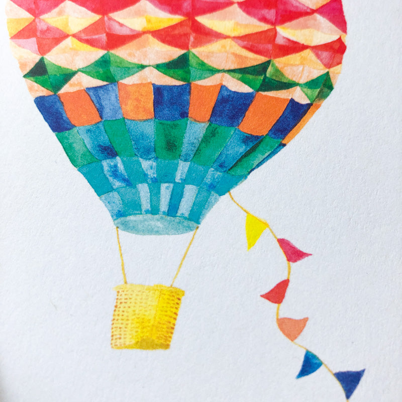 Colorful watercolor illustration of a vibrant hot air balloon with geometric patterns, featuring a yellow basket and trailing multicolored flags, perfect for a graduation celebration card