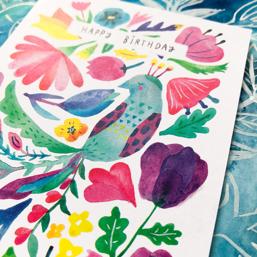 Colorful watercolor birthday card featuring a stylized peacock surrounded by vibrant flowers and foliage, with "Happy Birthday" text in the center, displayed on a blue patterned background