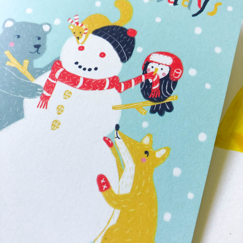 Cheerful holiday greeting card featuring a smiling snowman with a red scarf, accompanied by a penguin, fox, and polar bear amid falling snow on a light blue background