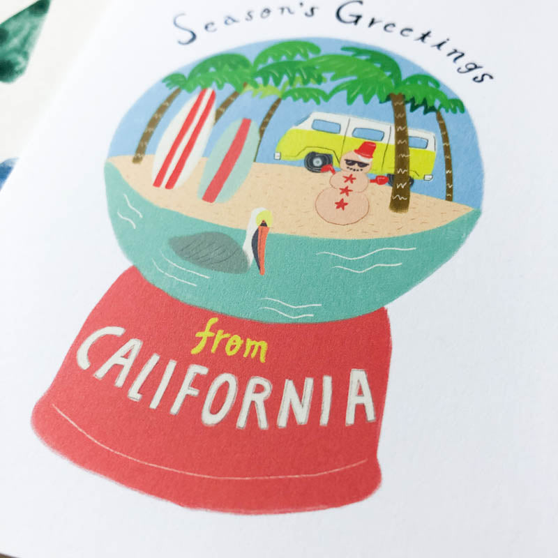 Snow globe featuring California beach scene with palm trees, surfboards, yellow van, and sandman, on red base with "from CALIFORNIA" text. "Season's Greetings" at top of globe.