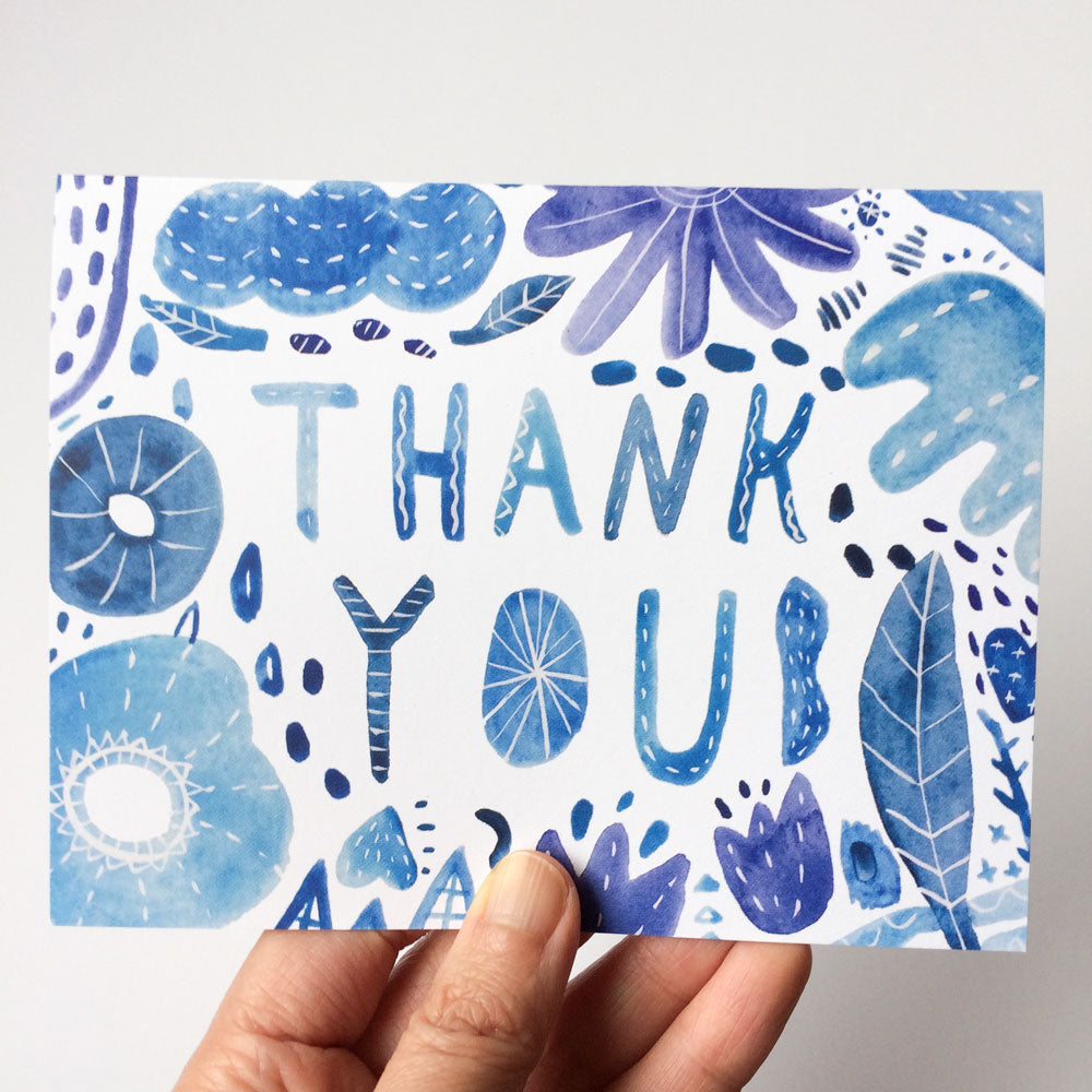 Handmade watercolor thank you card in shades of blue with floral and leaf designs, featuring "THANK YOU" text in artistic lettering, held by a hand against a white background