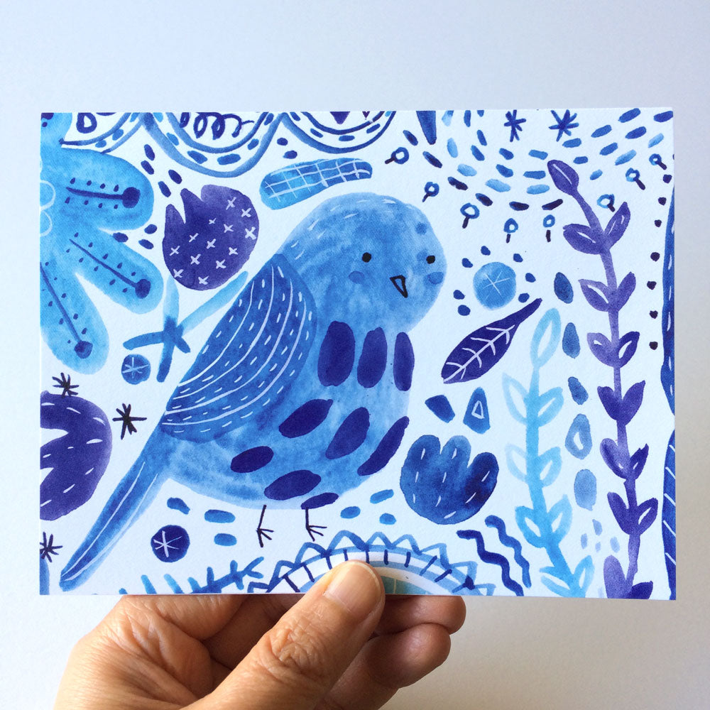 Watercolor illustration of a blue bird surrounded by whimsical floral and abstract elements in shades of blue and purple, printed on a notecard held by a hand