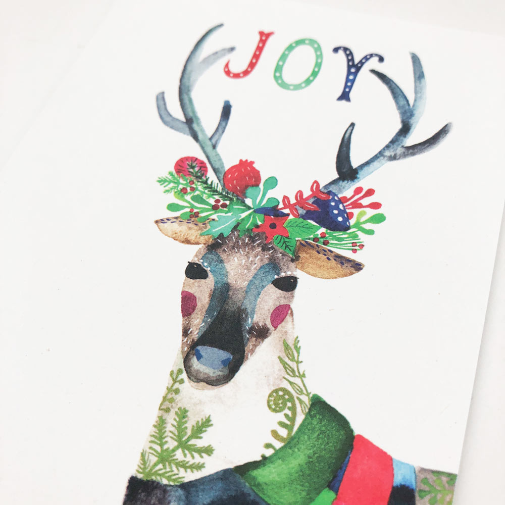 Watercolor illustration of a festive reindeer wearing a colorful scarf, with antlers spelling 'JOY' and adorned with holiday florals, berries, and greenery
