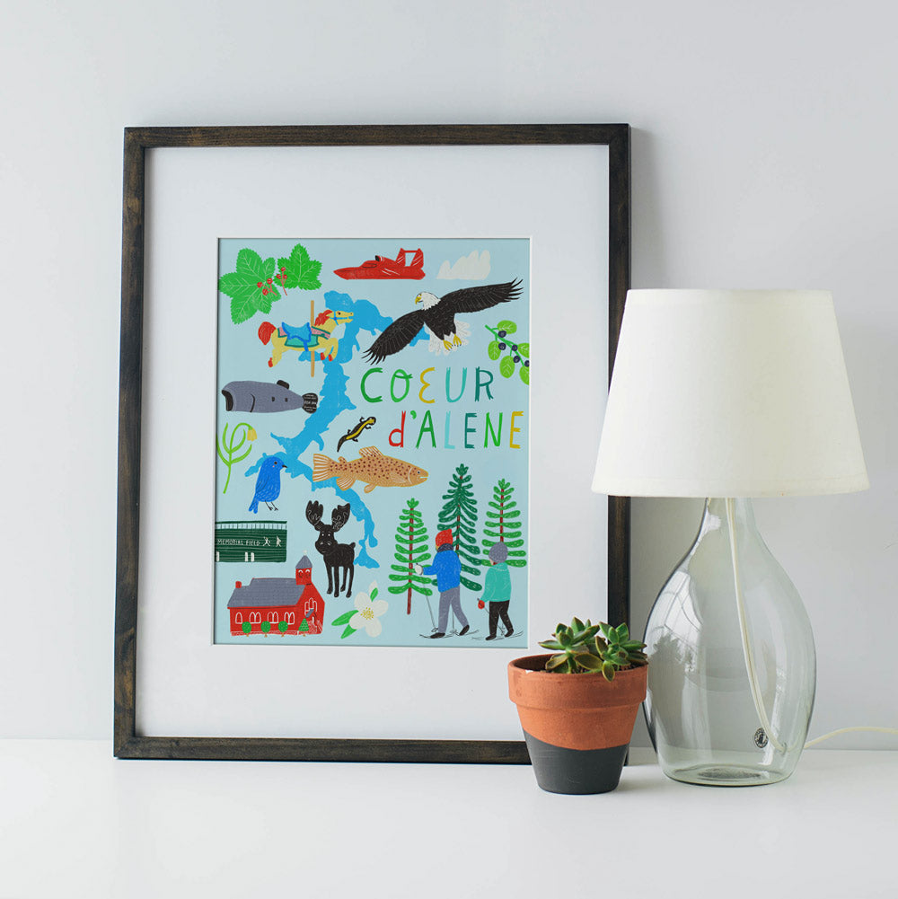Colorful illustrated giclee art print of Coeur d'Alene, Idaho, featuring local landmarks, wildlife, and activities, framed and displayed next to a lamp and plant pot