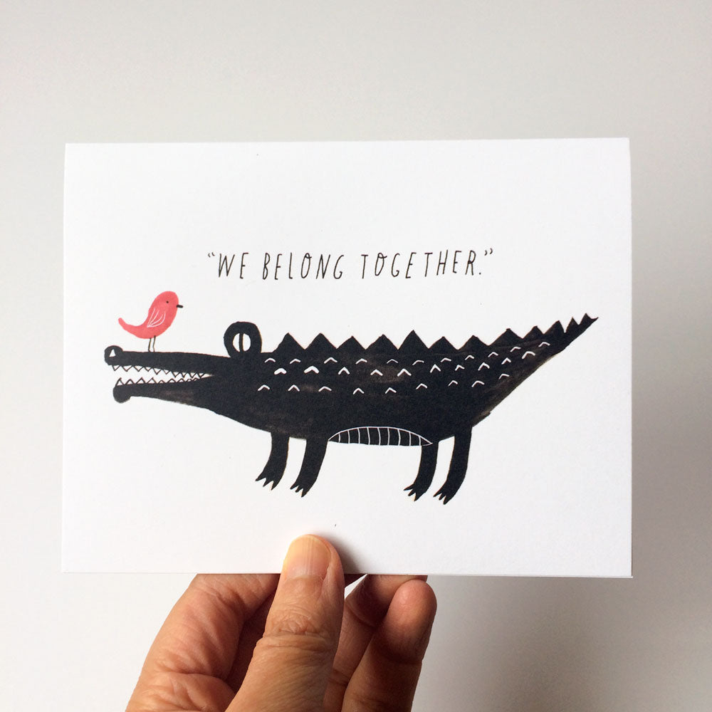 Watercolor illustration of a black crocodile with a pink bird perched on its snout, captioned "We Belong Together" on a white card held by a hand, perfect for wedding or anniversary greetings.