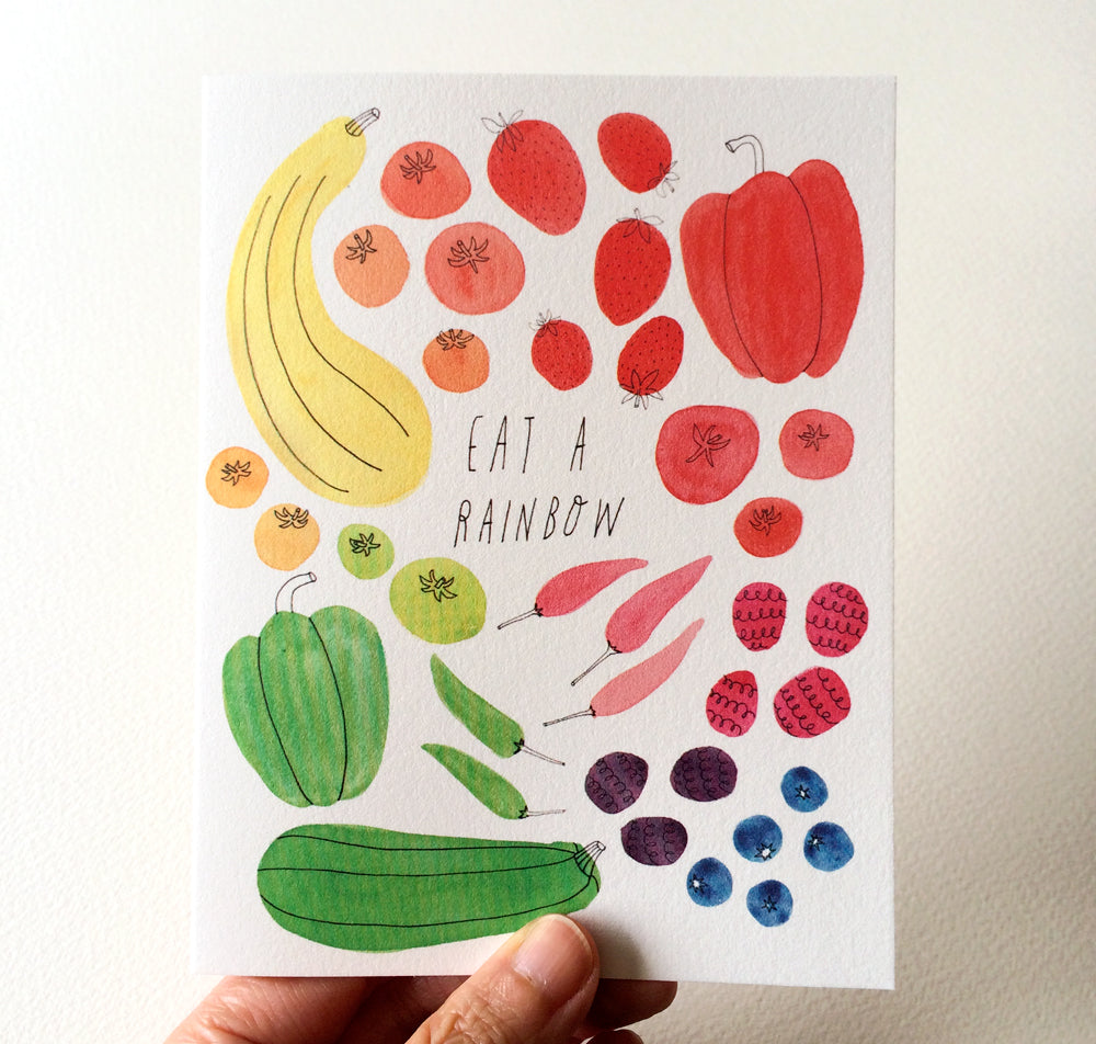 Colorful watercolor illustration of fruits and vegetables arranged in a rainbow pattern on a greeting card, with "Eat a Rainbow" text in the center. Hand holding the card visible at the bottom.