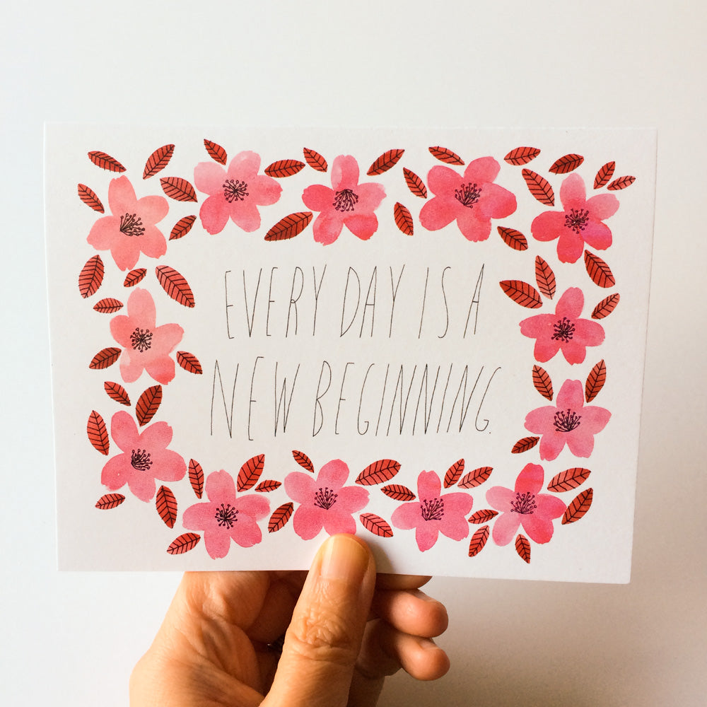 Greeting card with hand-drawn pink flowers and leaves framing the handwritten text "Every day is a new beginning" held by a hand against a white background