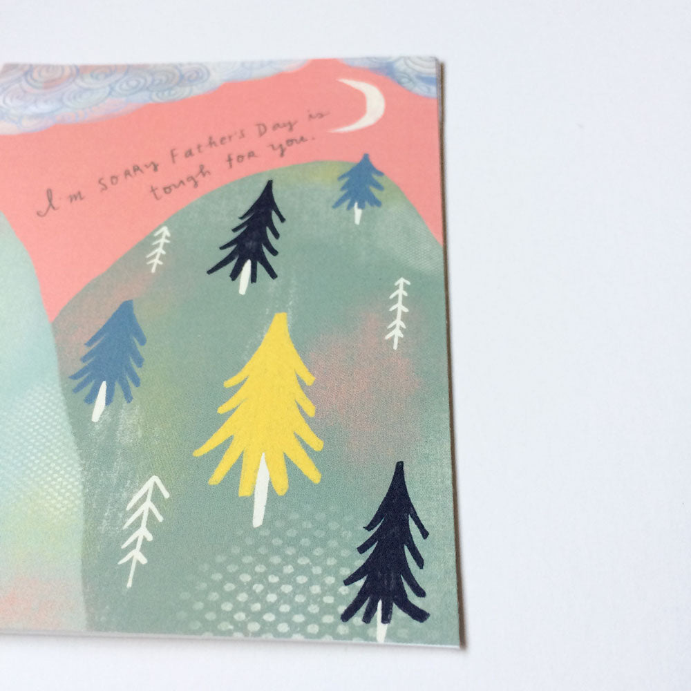 Father's Day empathy card featuring a whimsical illustration of mountains with colorful trees under a crescent moon, with handwritten text "I'm sorry Father's Day is tough for you" on a pink sky background.