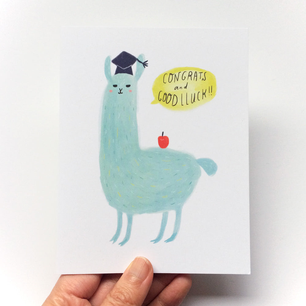 Cute illustrated graduation card featuring a light blue llama wearing a graduation cap, with a speech bubble saying "Congrats and Good Luck" and a small red apple, held by a hand against a white background