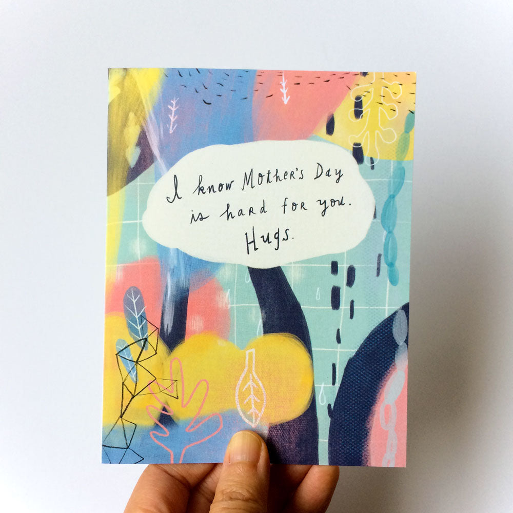 Mother's Day empathy card with colorful abstract background and handwritten message "I know Mother's Day is hard for you. Hugs." Card held by hand against white surface.