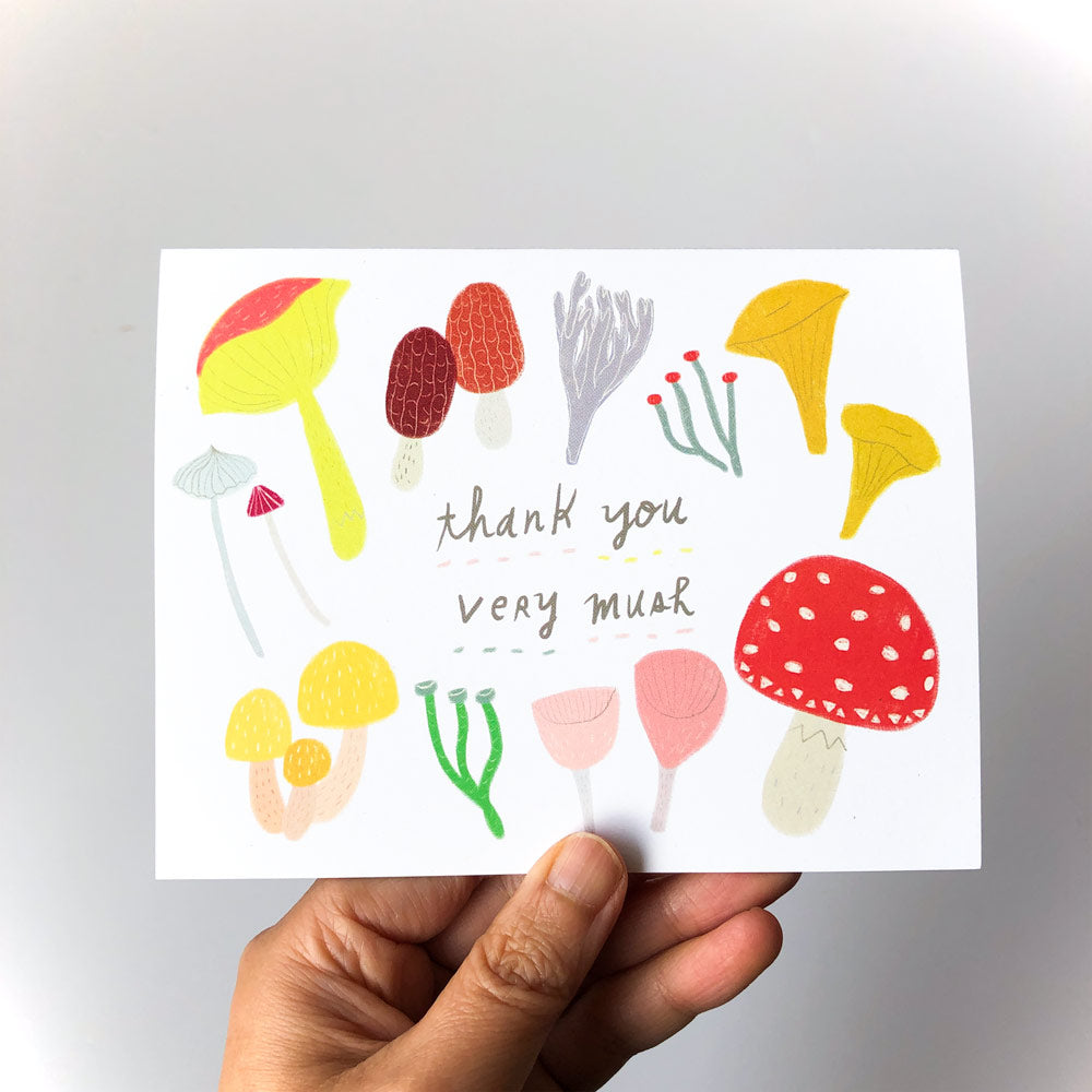 Hand holding a colorful greeting card featuring various illustrated mushrooms and fungi with the text "thank you very mush" in handwritten style, showcasing a whimsical and nature-inspired design for expressing gratitude.