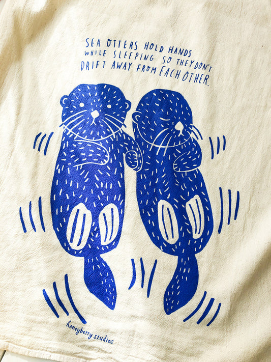 Flour sack tea towel with blue screen-printed design of two sea otters holding hands, with text explaining they do this while sleeping to avoid drifting apart