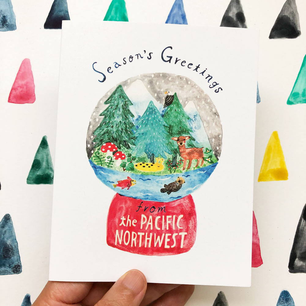 Watercolor greeting card featuring a snow globe with Pacific Northwest scene, including pine trees, wildlife, and colorful mushrooms. The globe sits on a red base labeled "from the PACIFIC NORTHWEST". Surrounded by vibrant triangle shapes, with "Season's Greetings" written above.