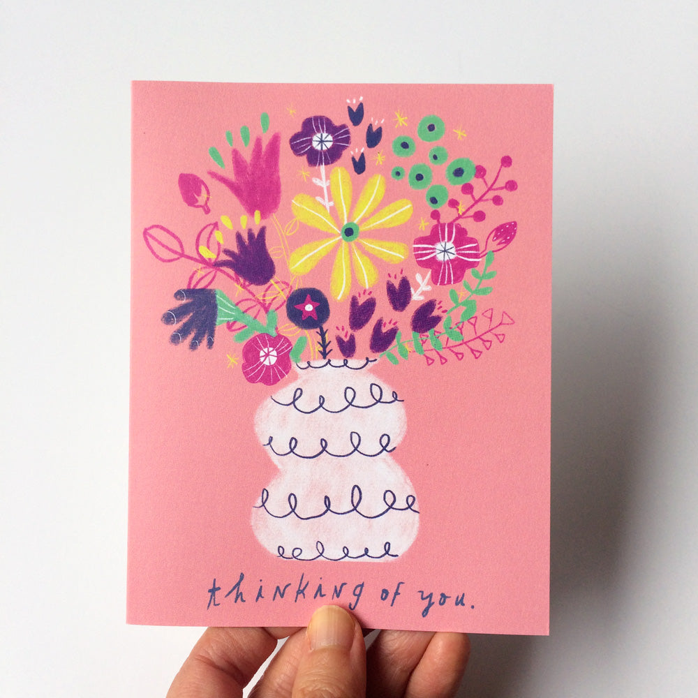 Colorful hand-drawn floral bouquet in whimsical vase on pink greeting card, featuring vibrant purple, yellow, and green flowers with "thinking of you" message, held against light background