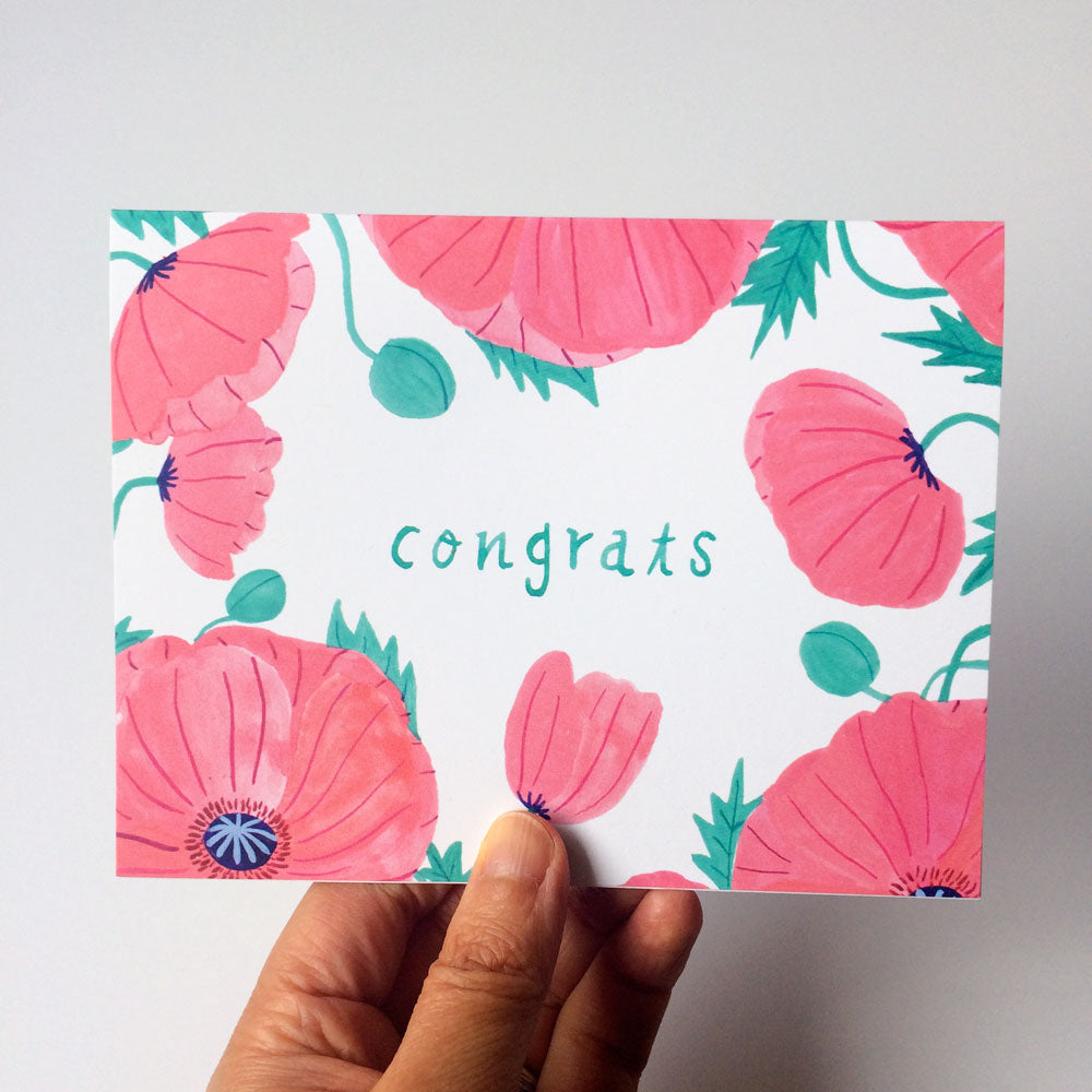 Watercolor greeting card featuring pink oriental poppies with teal leaves and stems, with "congrats" written in teal cursive font in the center, held by a hand against a light background