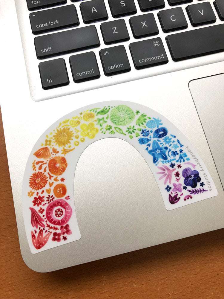 Colorful rainbow arch sticker featuring various flowers and natural elements in vibrant hues, placed on a laptop keyboard. The sticker showcases a gradient of colors from red to purple, creating a cheerful and diverse floral design.
