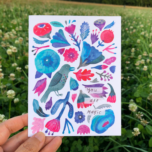 Colorful watercolor greeting card featuring whimsical floral and bird designs in vibrant blues, pinks, and purples, with "You are Magic" text, held against a grassy field background