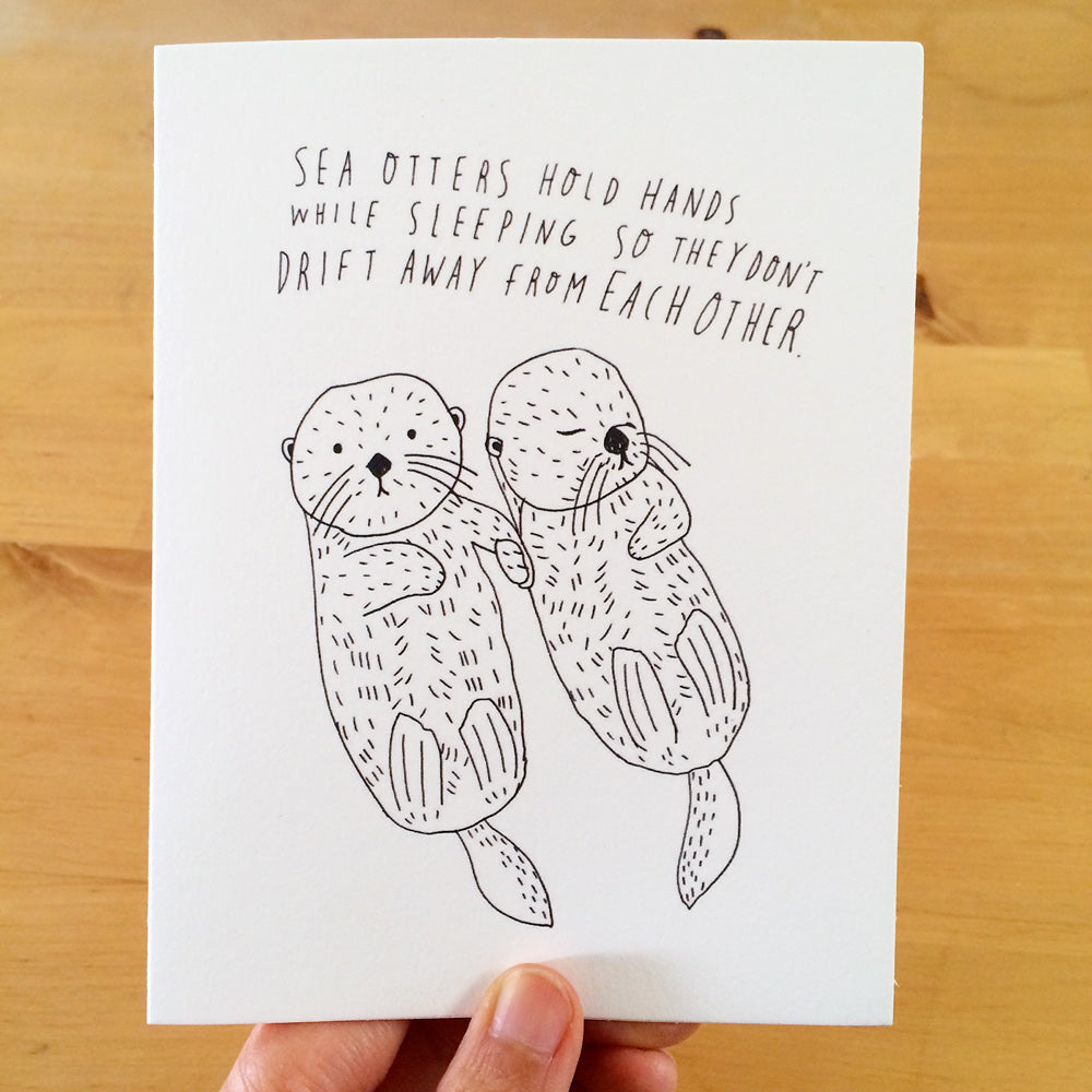 Hand-drawn greeting card featuring two adorable sea otters holding hands while sleeping, with handwritten text explaining their behavior to prevent drifting apart. Simple black ink illustration on white card stock.