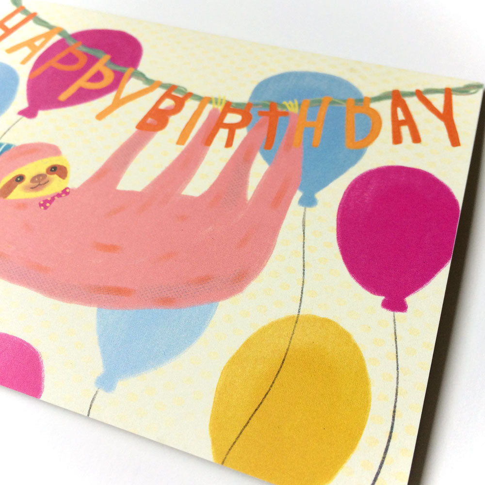 Colorful birthday card featuring a cartoon sloth hanging from a 'Happy Birthday' banner, surrounded by vibrant balloons in pink, blue, and yellow, creating a cheerful and whimsical design