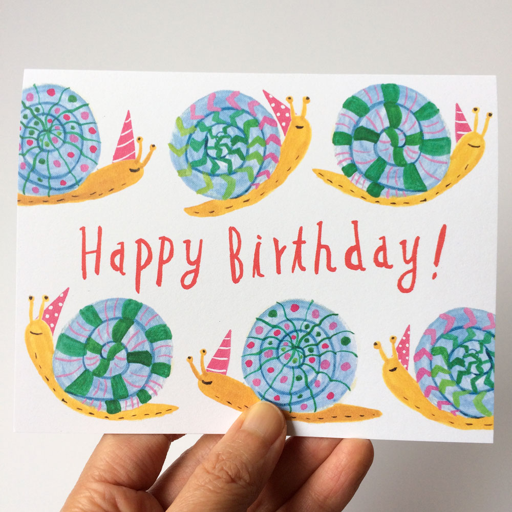 Colorful birthday card featuring six cartoon snails with patterned shells in blue, green, and purple, wearing party hats. Text reads "Happy Birthday!" in red. Hand holding the card visible at bottom.