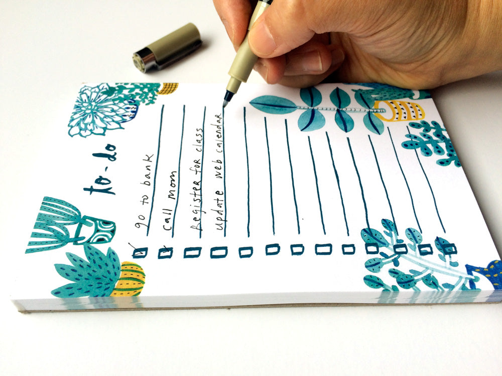 Hand writing on a decorative to-do list notepad featuring watercolor succulent illustrations in blue and green hues, with lined spaces and checkboxes for task organization