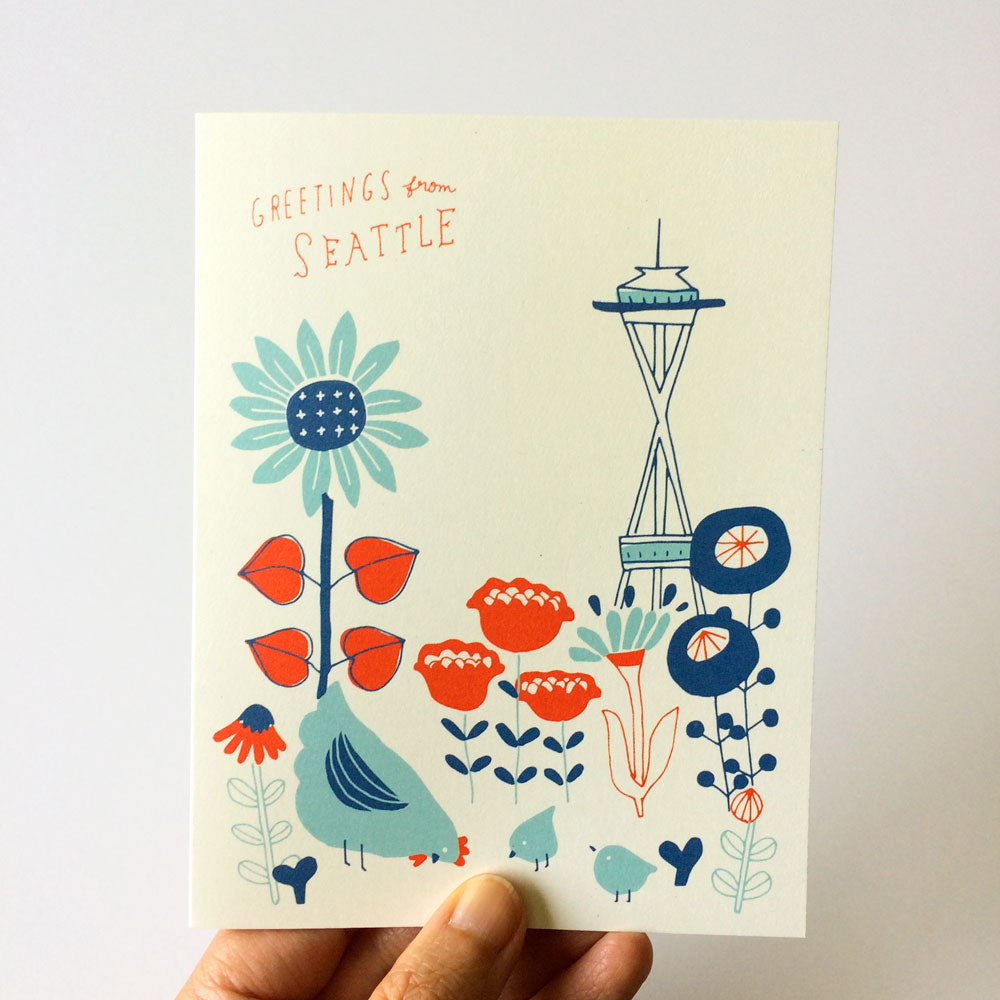Vintage-style Seattle greeting card featuring stylized flowers, chickens, and Space Needle against a pale background. Hand-drawn text reads "Greetings from Seattle" in coral.