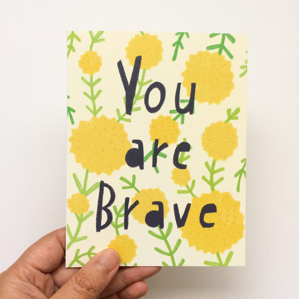 Hand holding a card with "You are Brave" text surrounded by yellow dandelion-like flowers and green leaves on a cream background, perfect for encouragement and support.