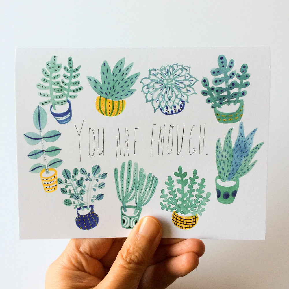 Watercolor illustration of various potted plants and succulents surrounding handwritten text "You are enough" on a white card held by a hand, showcasing a motivational greeting card design