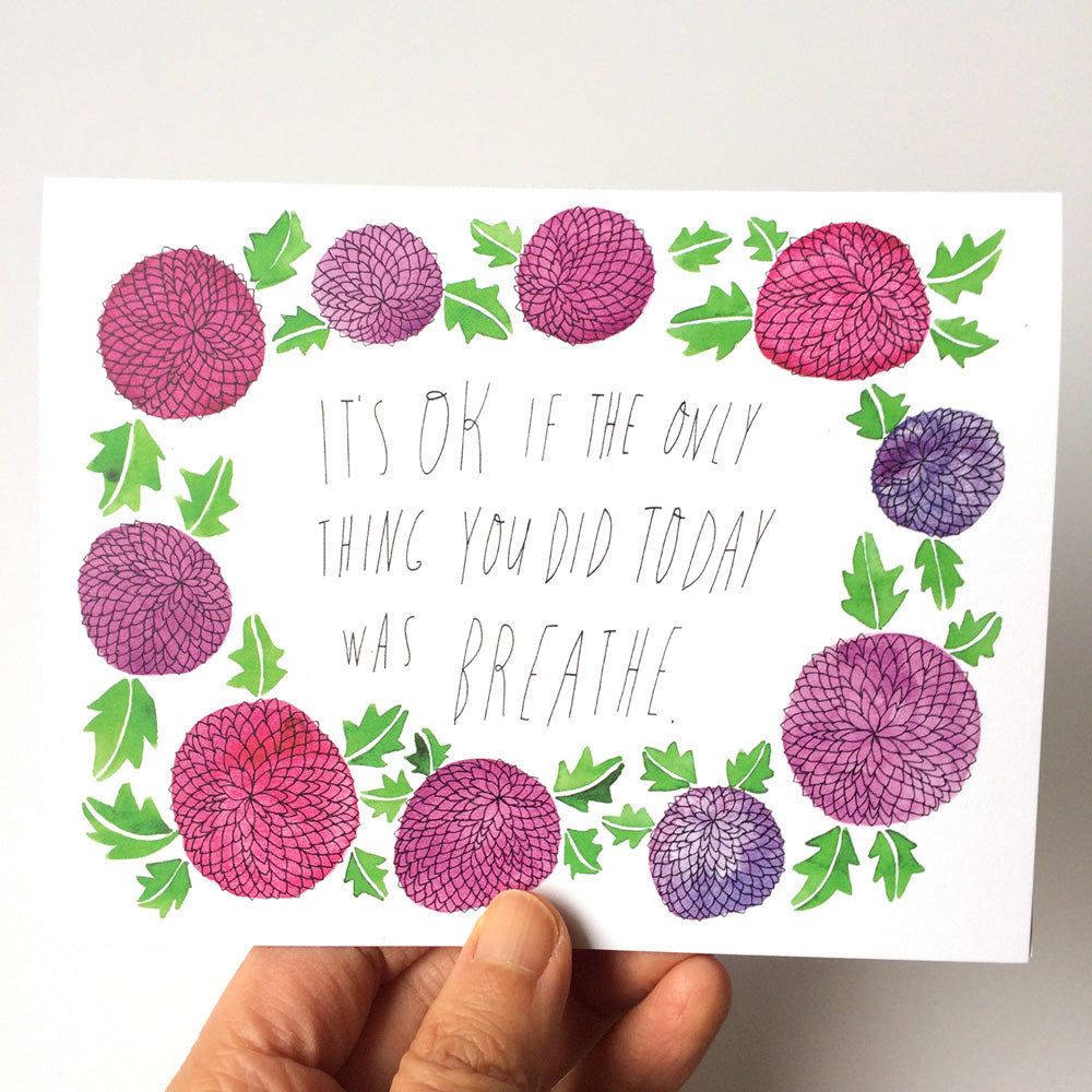 Watercolor greeting card with pink and purple flower illustrations surrounding handwritten text "It's OK if the only thing you did today was breathe", held by a hand against a white background.