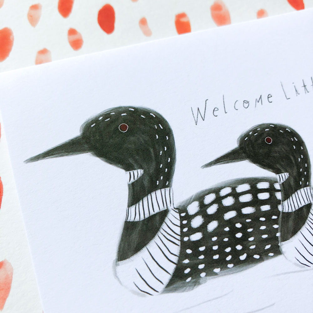 Hand-drawn loon greeting card with two black and white loons, featuring stylized patterns on their bodies. Text reads "Welcome Lil" with coral-colored abstract shapes bordering the top and left edges of the card.