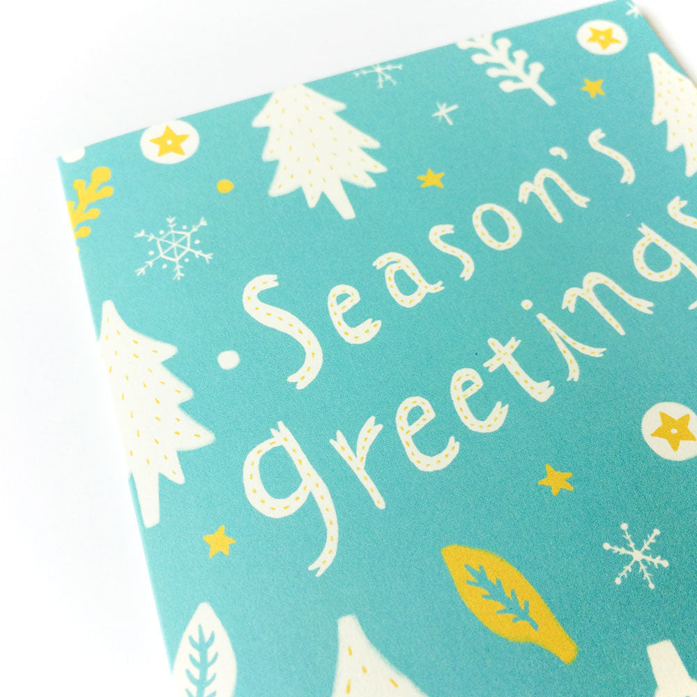 Teal holiday greeting card with "Season's greetings" text, decorated with white snowflakes, Christmas trees, yellow stars, and leaf patterns, showcasing a festive winter design for holiday wishes
