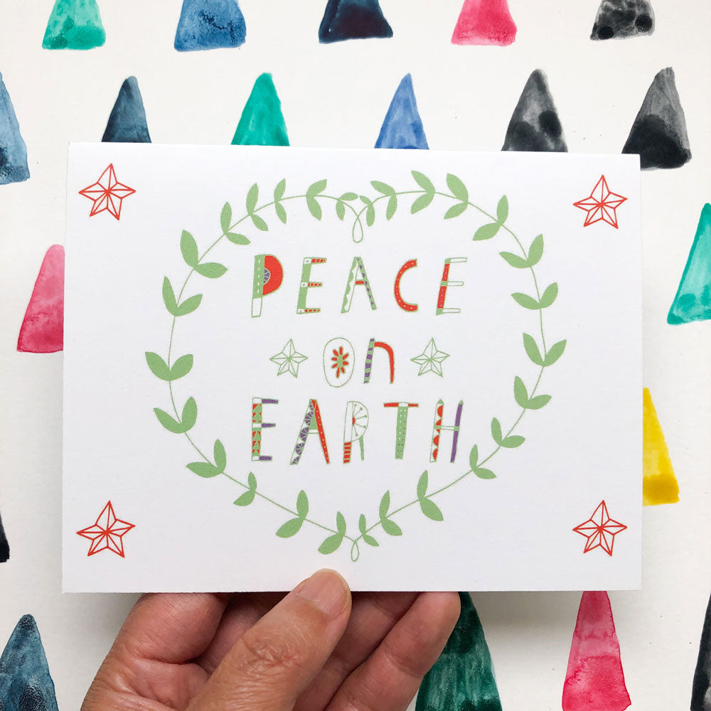 Hand-lettered holiday card with "Peace on Earth" text in colorful letters, surrounded by green wreath and stars, held against background of watercolor triangles in various colors