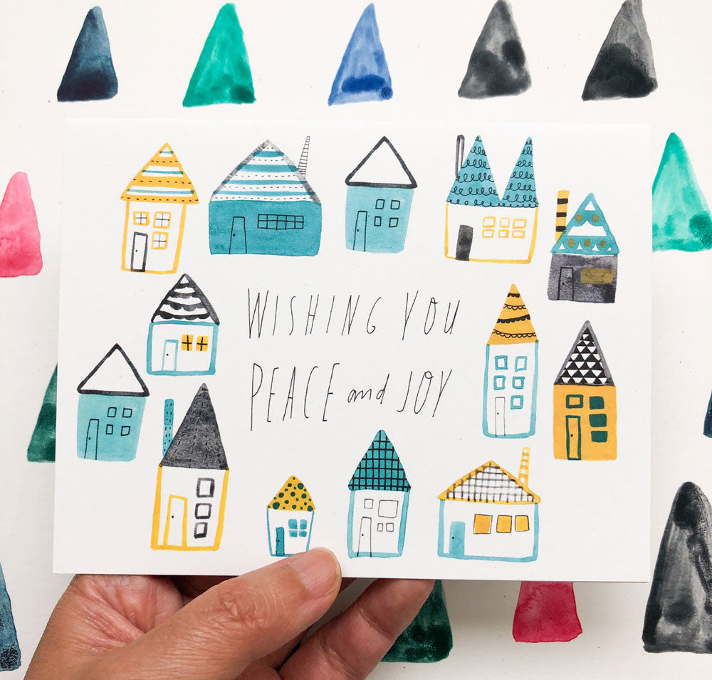 Watercolor holiday greeting card featuring whimsical folk houses in teal and yellow with handwritten message "Wishing You Peace and Joy" held against colorful triangular background