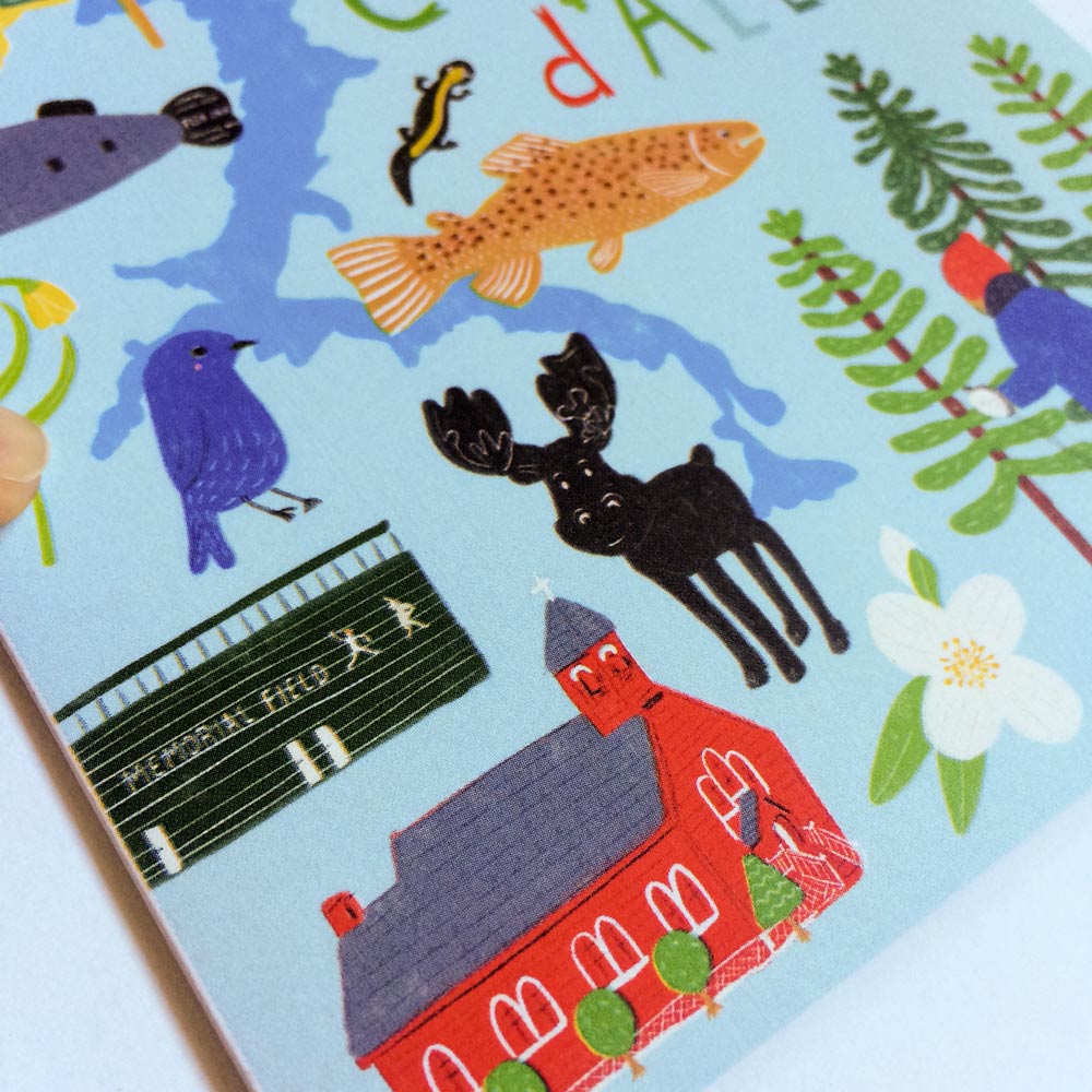 Colorful illustrated greeting card featuring Coeur d'Alene landmarks and wildlife, including a fish, bird, moose, and red building, surrounded by nature elements on a light blue background with hand-lettered text.