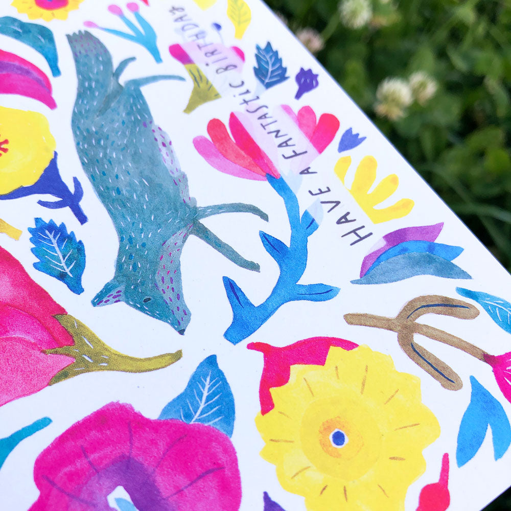 Colorful watercolor birthday card featuring a blue fox silhouette surrounded by vibrant flowers, leaves, and the text 'Have a fantastic' in whimsical lettering, set against a white background