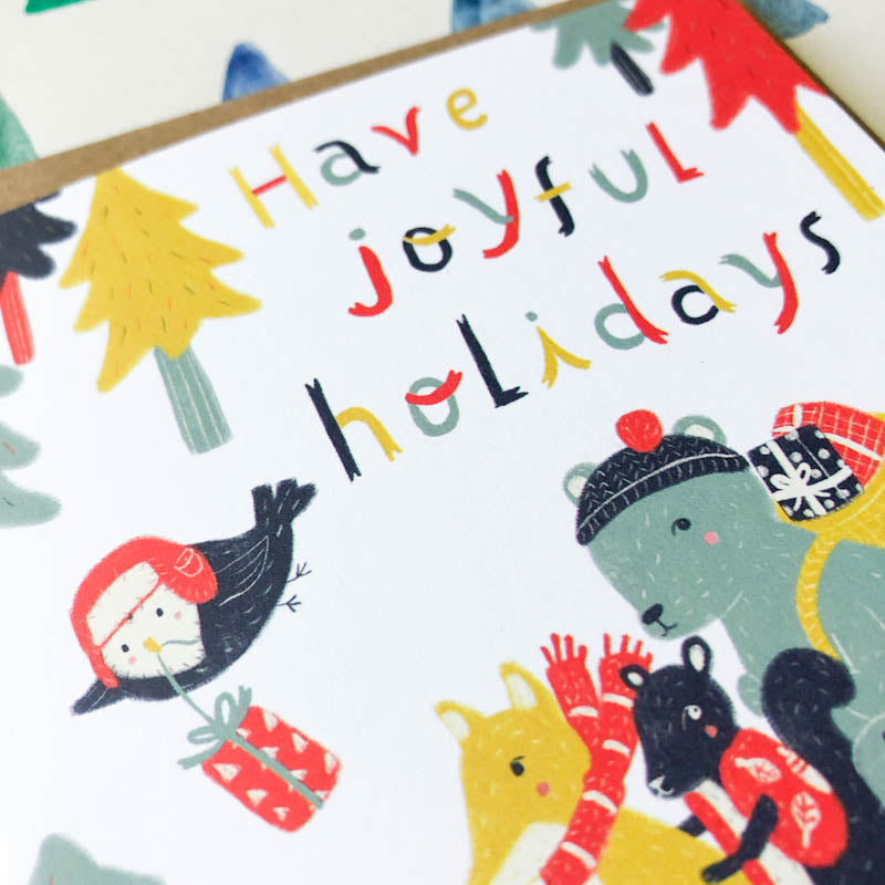Colorful holiday greeting card featuring whimsical woodland animals in winter attire, with playful text "Have joyful holidays" surrounded by festive trees and a gift, showcasing a cheerful and cute design for seasonal greetings.