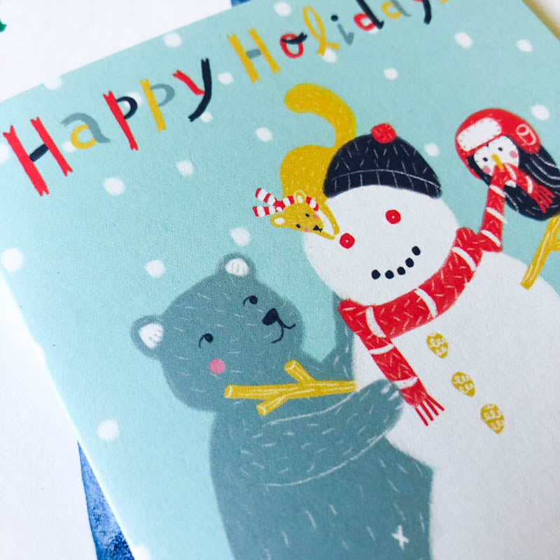 Cheerful holiday greeting card featuring a cute snowman wearing a hat and scarf, accompanied by a smiling gray bear, with colorful "Happy Holiday" text on a light blue background with falling snow