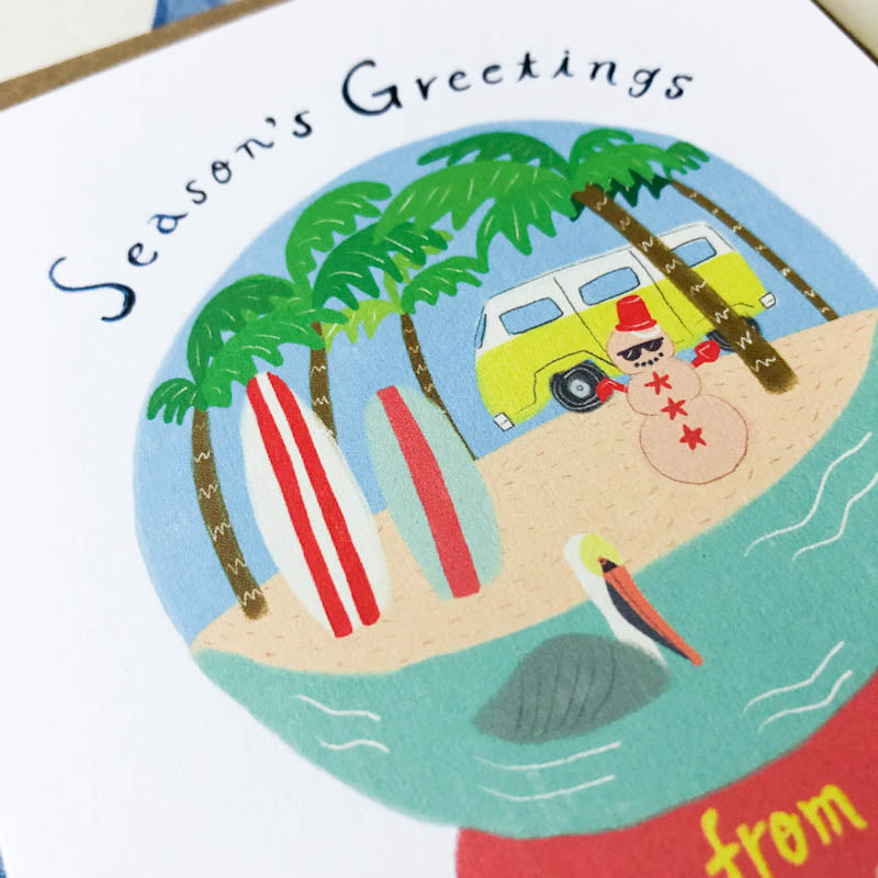 Season's Greetings holiday card featuring a beach scene snow globe with palm trees, yellow van, surfboards, and snowman on sand, surrounded by blue water, evoking a Southern California Christmas vibe