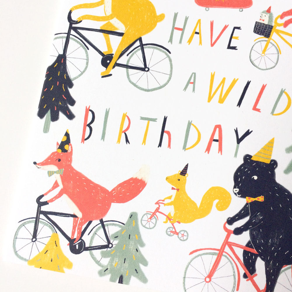 Whimsical birthday card design featuring woodland animals riding bicycles, with text "Have a Wild Birthday" in colorful, playful lettering. Animals include a fox, squirrel, and bear, surrounded by stylized trees and party hats.