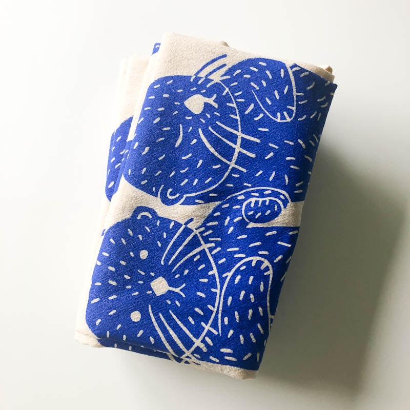 Folded flour sack tea towel with cobalt blue sea otter design, showing two otters holding hands, printed on natural unbleached cotton fabric. Slight imperfections visible, marketed as seconds quality.