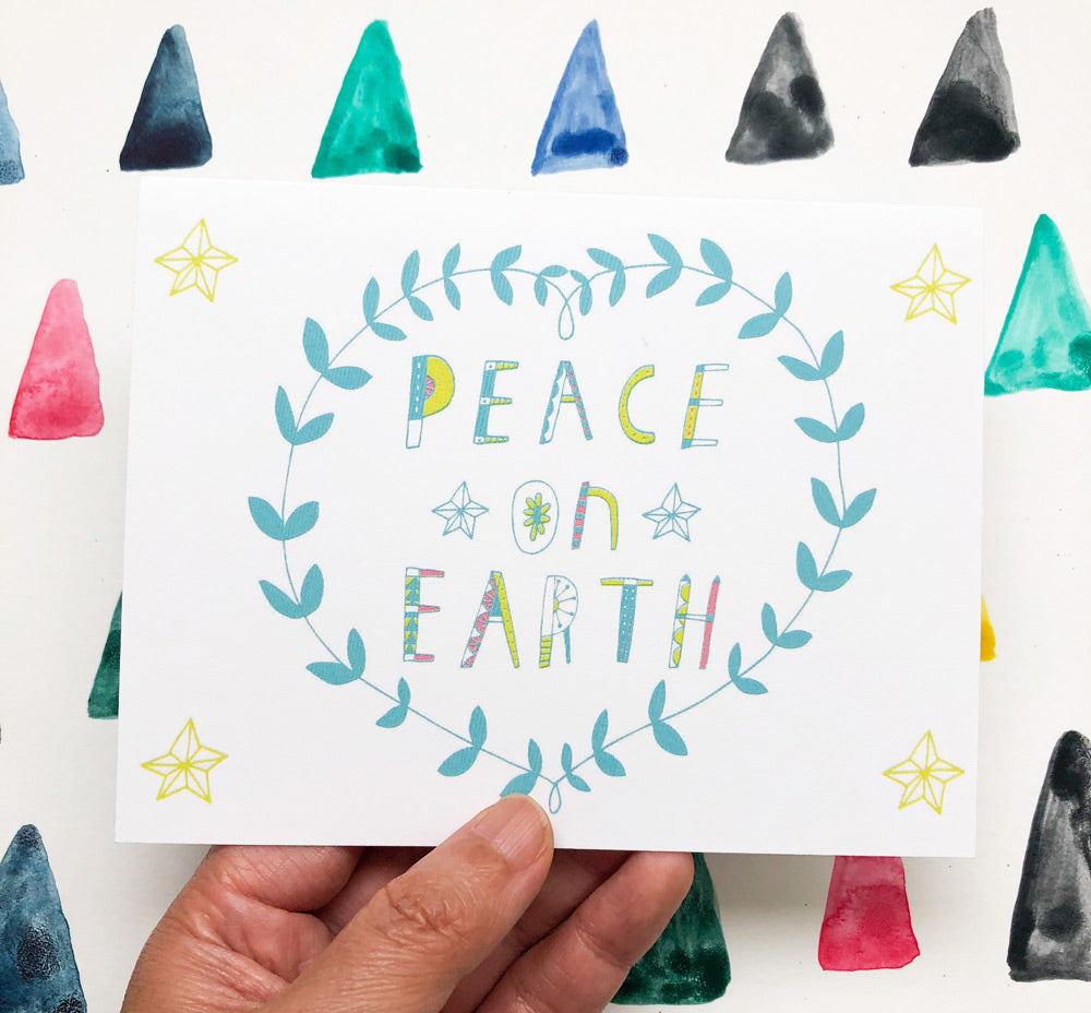 Hand-drawn holiday greeting card featuring "Peace on Earth" text in colorful letters, surrounded by a teal wreath and yellow stars, held against a background of watercolor triangles in various colors