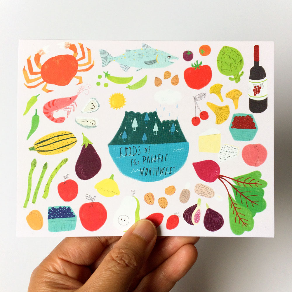 Colorful illustration of Pacific Northwest foods including seafood, fruits, vegetables, mushrooms, and wine, centered around a blue mountain labeled "Foods of the Pacific Northwest" on a white notecard held by a hand