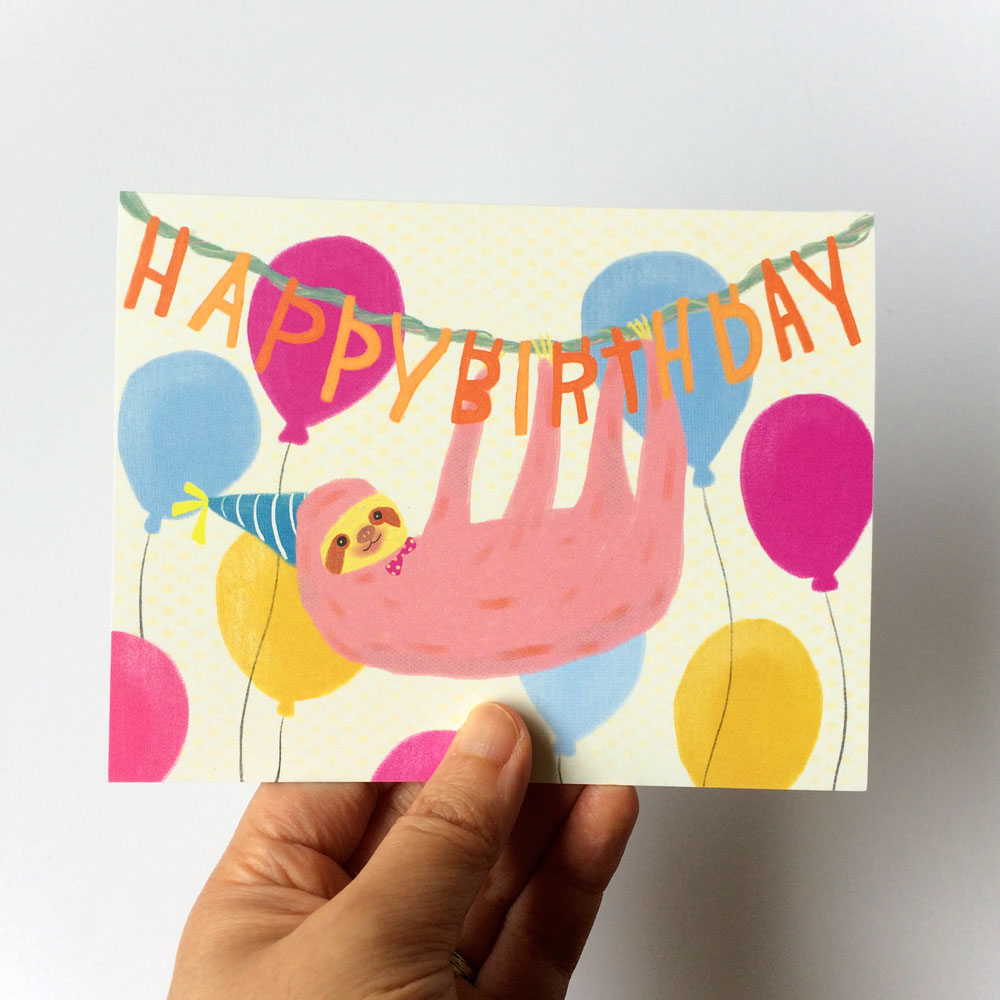 Colorful birthday card featuring a smiling pink sloth wearing a party hat, hanging from a "Happy Birthday" banner surrounded by festive balloons in blue, yellow, and magenta, held by a hand against a white background.