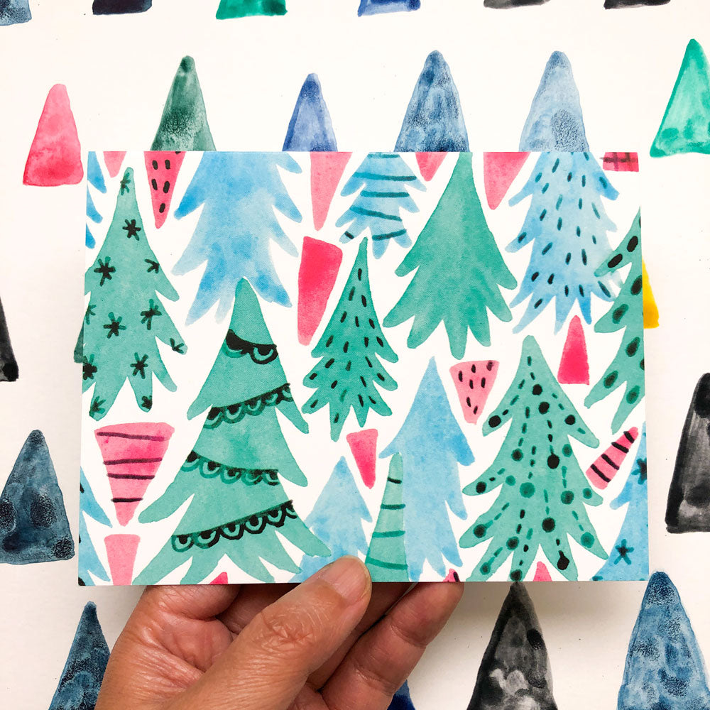 Whimsical watercolor illustration of a winter forest with stylized pine trees in various shades of green, blue, and pink, creating a festive and colorful holiday scene on a greeting card held by a hand