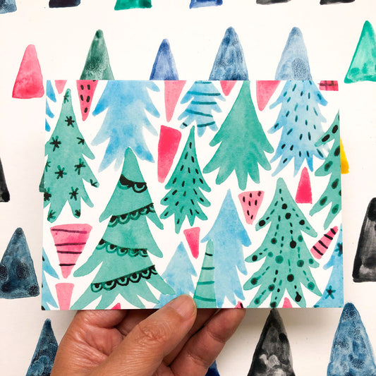 Whimsical watercolor illustration of a winter forest with stylized pine trees in various shades of green, blue, and pink, creating a festive and colorful holiday scene on a greeting card held by a hand