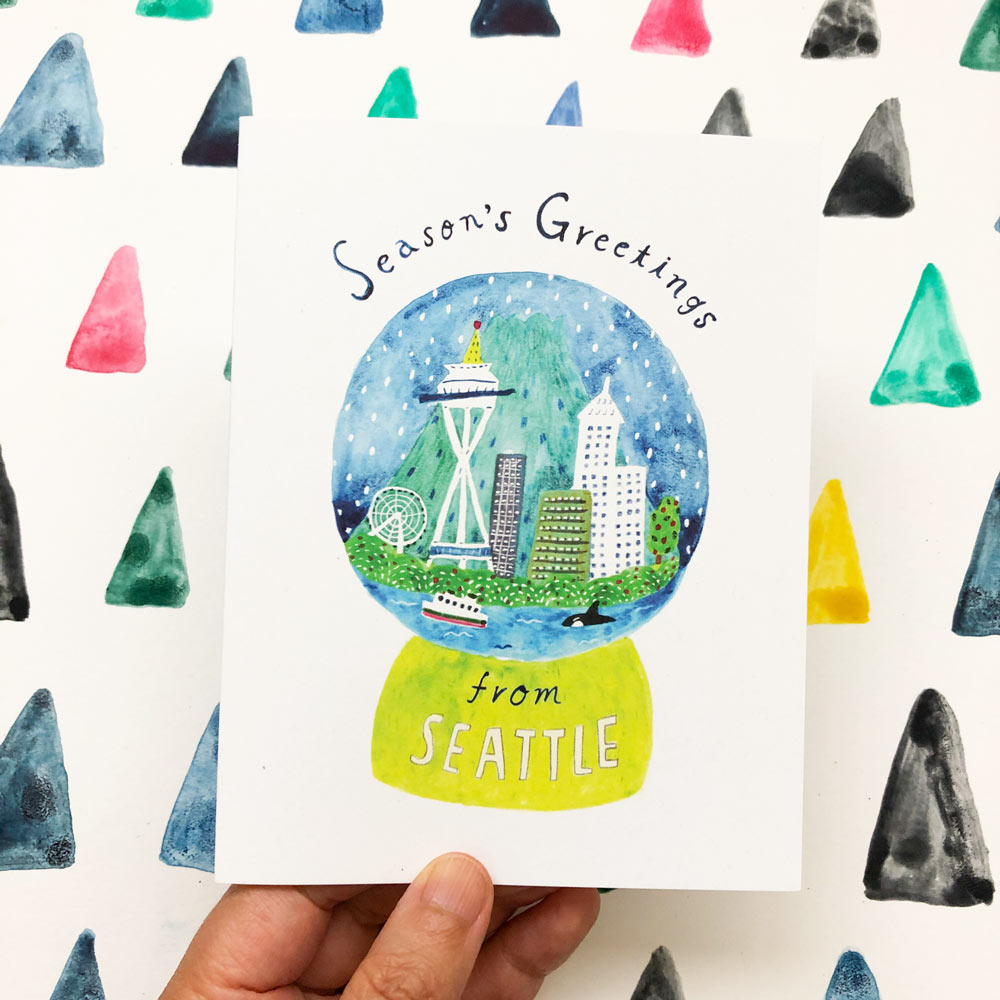 Watercolor greeting card featuring a Seattle snow globe with iconic landmarks, surrounded by colorful triangles. Hand holding the card, showcasing "Season's Greetings from Seattle" text and vibrant cityscape inside the globe.